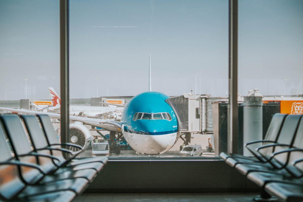 A photo looking out from an airport terminal window. The nose of the blue plane is prominent. Chairs are on both sides of the terminal window.